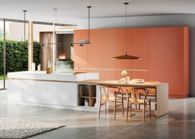 Cocina blanca e isla con muebles columna terracota, mesa de madera con sillas Wishbone y grandes ventanales al jardín