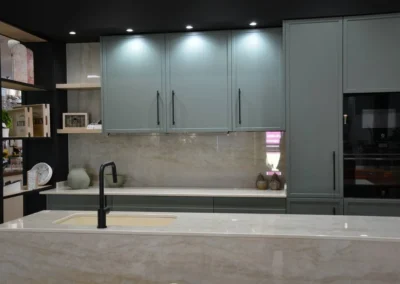 Detalle de cocina verde salvia con isla de encimera de porcelánico, grifo negro y muebles altos en exposición Fuoco